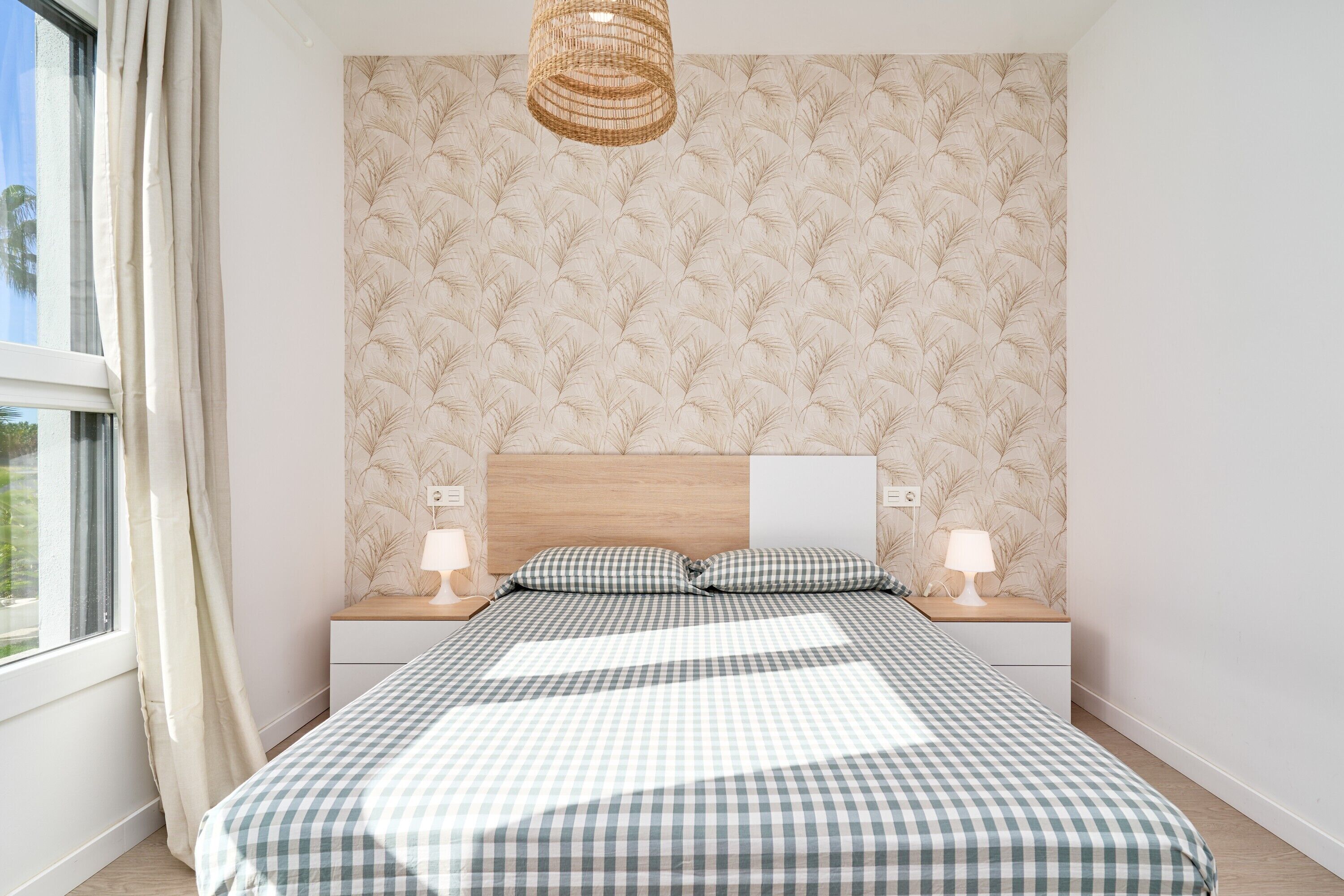 A bedroom with a checkered bedspread, a wooden headboard, bedside lamps, and a window with curtains.