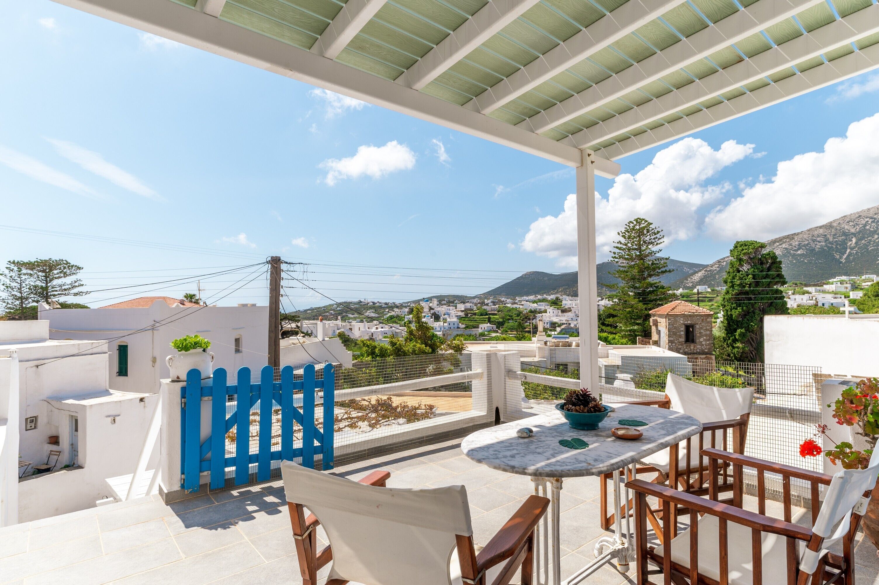 Una terraza con una mesa y sillas blancas, un portón azul y vistas a un pueblo y montañas.