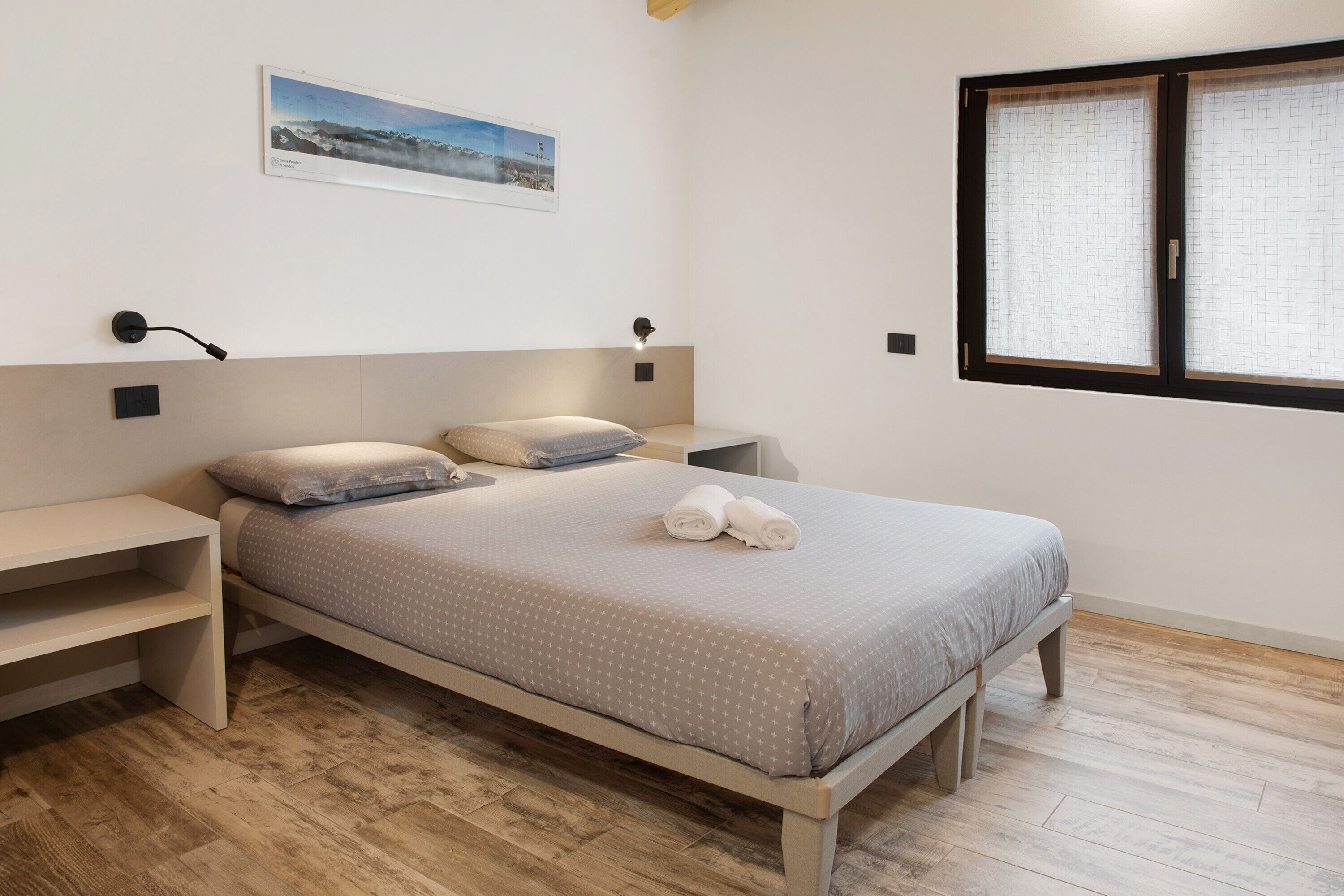 A bedroom with a bed, bedside tables, a wall-mounted lamp, a window with blinds, and a framed picture on the wall.