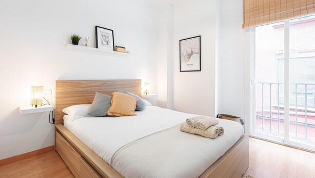 Een net opgemaakt bed met kussens, een bedlampje, een klein plankje met decoratieve spullen en een ingelijst beeld aan de muur.