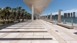 Een promenade aan het water met een moderne overkapping, palmbomen en een stadsachtergrond.