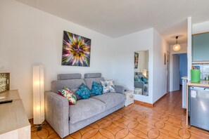 A living room with a grey sofa, colorful pillows, a modern abstract painting, a wooden cabinet, and a kitchen area with a fridge.
