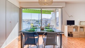 Un comedor con una mesa dispuesta para dos, un televisor sobre un mueble y vista al puerto deportivo a través de amplias ventanas.