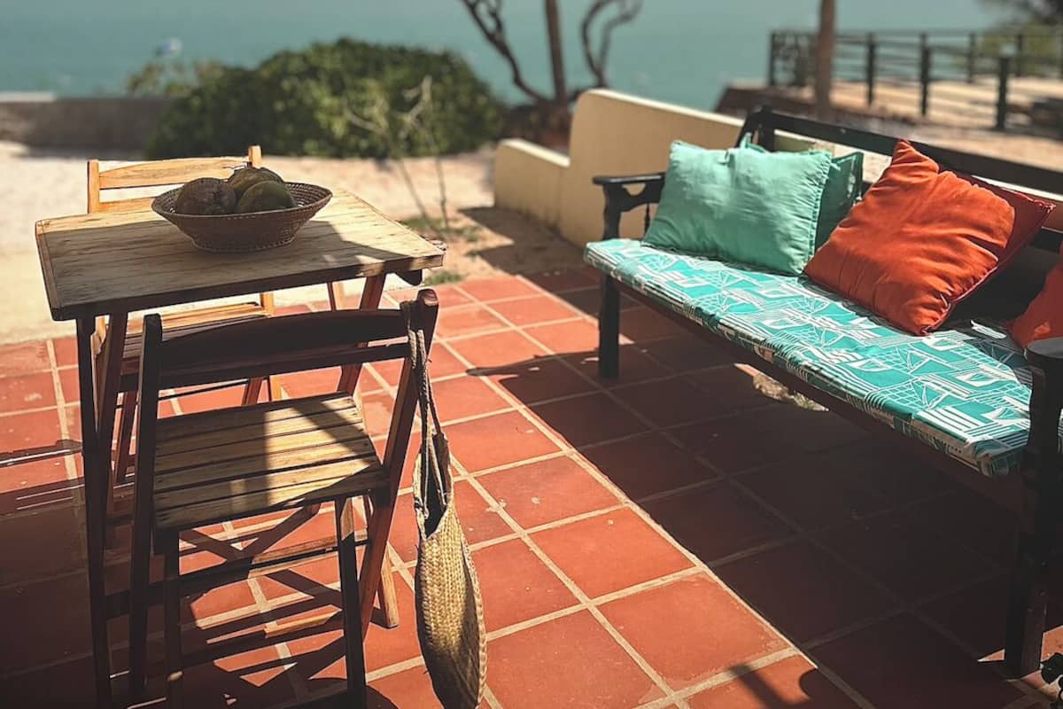 Un patio con una mesa, sillas y un sofá con cojines. Hay una cesta en la mesa y un bolso colgado en la silla. Palmeras y vistas al océano se observan al fondo.