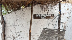 A hammock under a thatched roof, a white wall, and a sign reading "BARRAQUINHA RAIZ". - Pousada Vila Matury (Icapuí)
