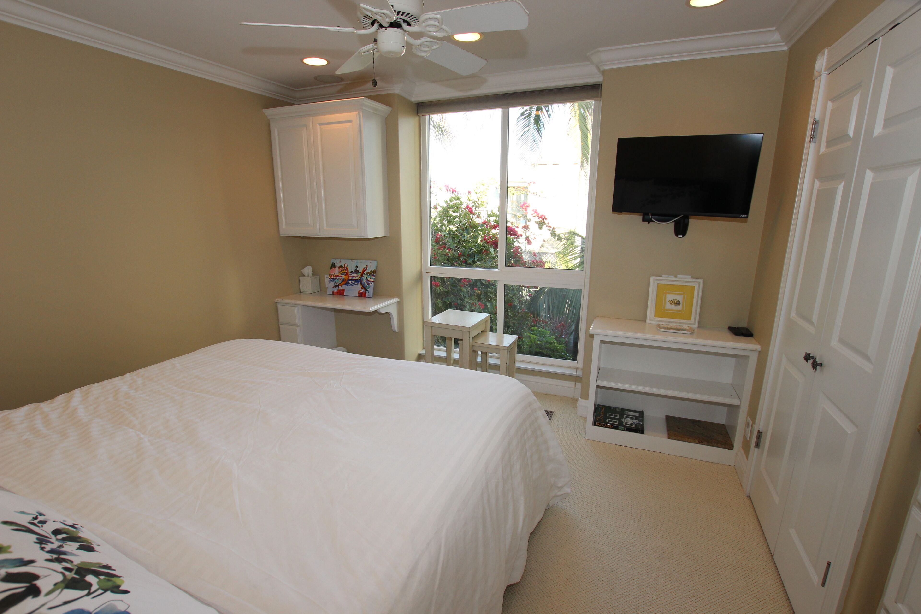 A bedroom with a bed, a ceiling fan, a TV mounted on the wall, and a window with a view of greenery.