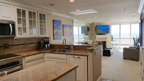 A modern kitchen with granite countertops, white cabinetry, and a large island. There is a dining area with a view of the ocean. - Tranquil 2 Bd Oceanfront Condo in charming Oceanside with WiFi, fitness room. (Oceanside)