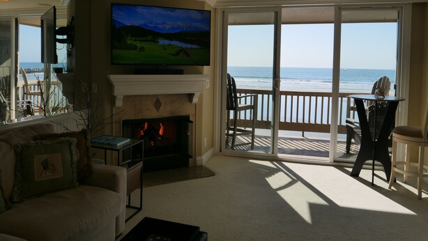 A living room with a fireplace, a television, and a view of the beach. - Tranquil 2 Bd Oceanfront Condo in charming Oceanside with WiFi, fitness room. (Oceanside)