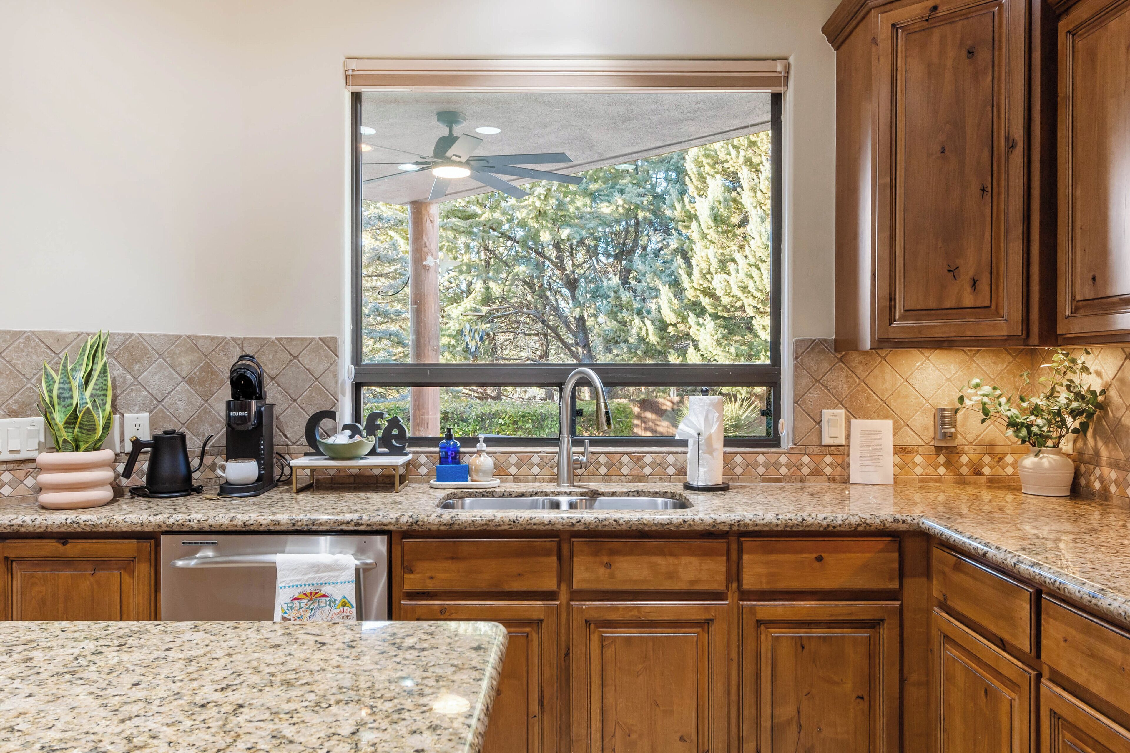 A kitchen with wooden cabinets, a granite countertop, a window with a view of trees, and a ceiling fan.