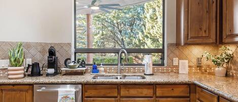 A kitchen with wooden cabinets, a granite countertop, a window with a view of trees, and a ceiling fan.