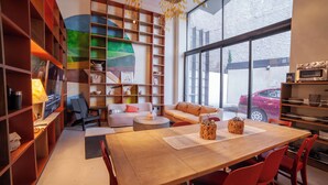 A modern living room with a wooden dining table, red chairs, a sofa, and a bookshelf filled with various items.