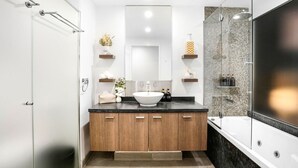 A modern bathroom with a large sink, wooden cabinets, a glass shower enclosure, and a mirror above the sink. - Luxury Studio in Palm Cove Resort | Steps to Beach & Pools (Palm Cove)