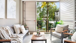 A modern living room with a sofa, armchairs, and a coffee table. There is a sliding glass door leading to a balcony with plants and a view of trees. - Luxury Studio in Palm Cove Resort | Steps to Beach & Pools (Palm Cove)