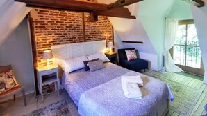 A bedroom with a sloped ceiling, exposed wooden beams, a brick wall, a bed with a headboard, bedside tables with lamps, and a window with curtains. - Charming self-catering cottage in a Plus Beau Village, near Pompadour. (Ségur-le-Château)