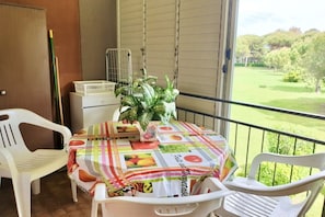 A balcony with a table covered in a fruit-themed tablecloth, white plastic chairs, and a potted plant.