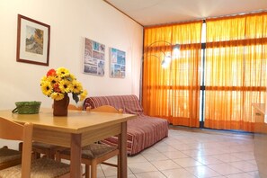A living room with a dining table, chairs, a sofa, and a large window with orange curtains.