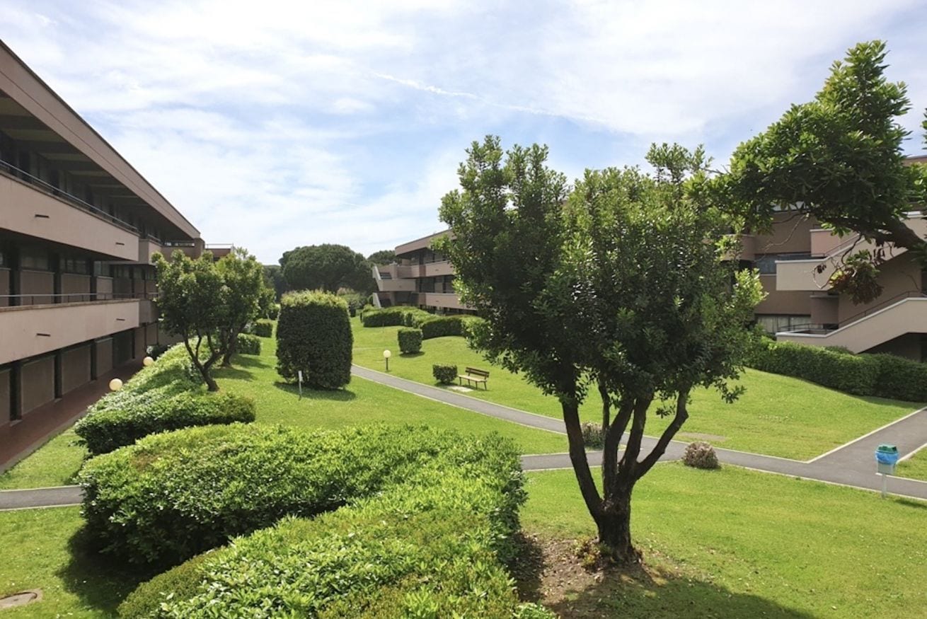 A courtyard with neatly trimmed hedges, a central tree, and modern buildings with balconies.