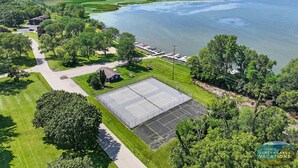 Vue aérienne d’une propriété au bord d’un lac, avec un court de tennis, un espace de stationnement et un petit bâtiment.
