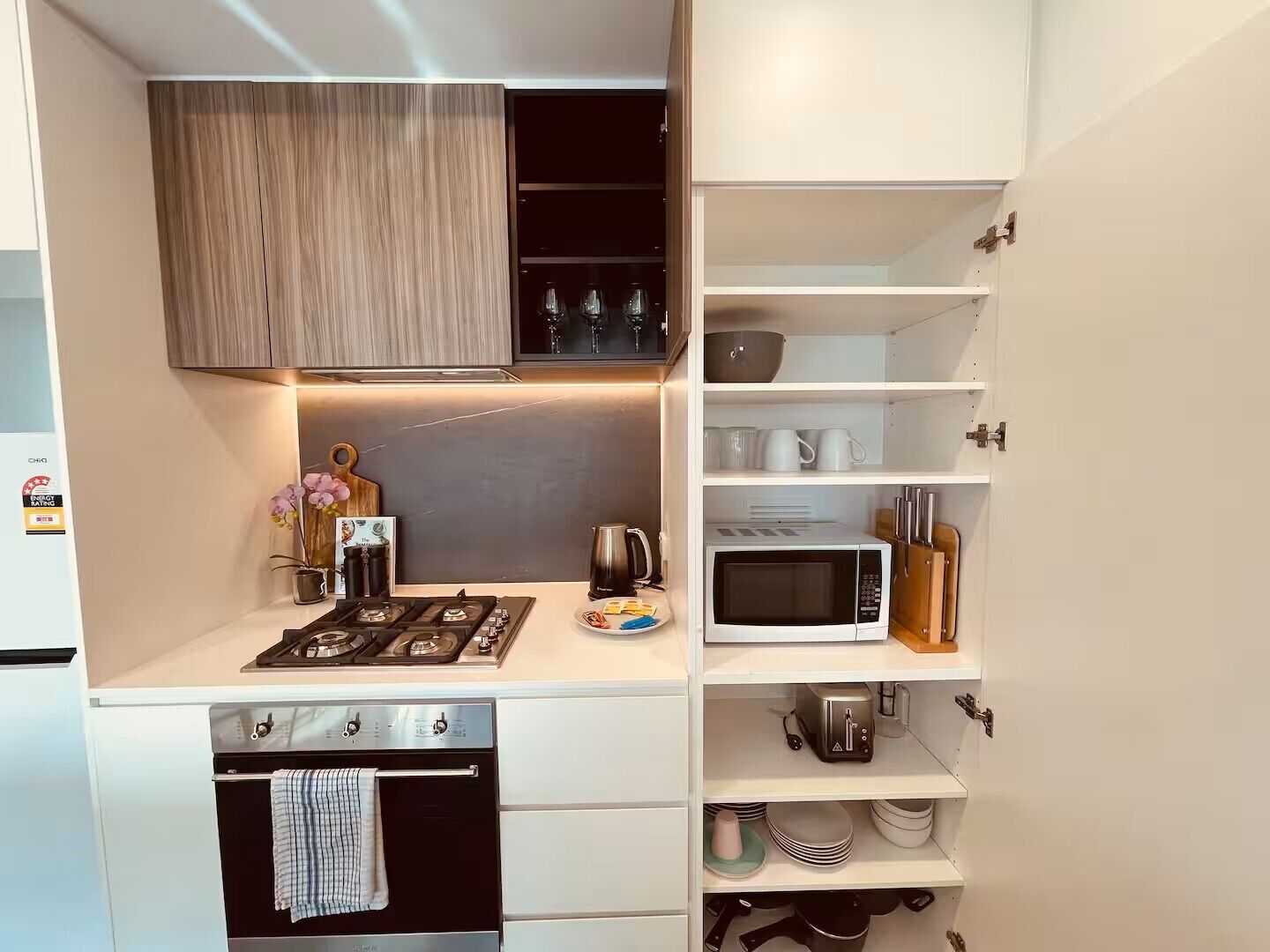 Uma cozinha compacta com fogão a gás, micro-ondas e forno. Há prateleiras com utensílios de cozinha e um balcão pequeno com uma cafeteira e um prato de comida.