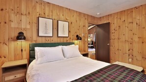 Una habitación con paneles de madera, una cama, dos cuadros enmarcados, una mesita de noche y una puerta que da a otra habitación.