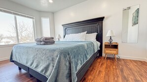 A bedroom with a large bed, wooden flooring, a nightstand with a lamp, and a window with blinds. - Spacious and conveniently located 5 Bedroom 3 Bath home in SLC with Amenities (Salt Lake City)