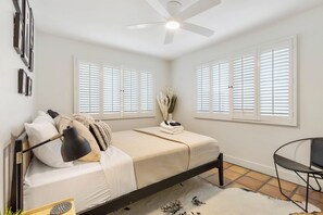 Un dormitorio con una cama, ventilador de techo, dos ventanas con contraventanas, una mesita y una silla.