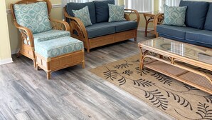 Ein Wohnzimmer mit Deckenventilator, zwei Korbschaukelsofas mit gemusterten Kissen, einem Glastisch und einem großen Fenster mit weißen Vorhängen.