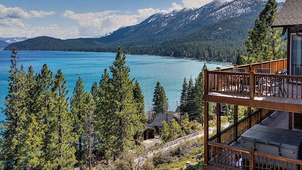 A mountain cabin with a deck overlooking a turquoise lake and snow-capped mountains.