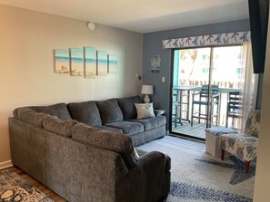 Ein Wohnzimmer mit grauem Ecksofa, einem kleinen Tisch mit Lampe und Blick auf die Außenpatio-Terrasse.