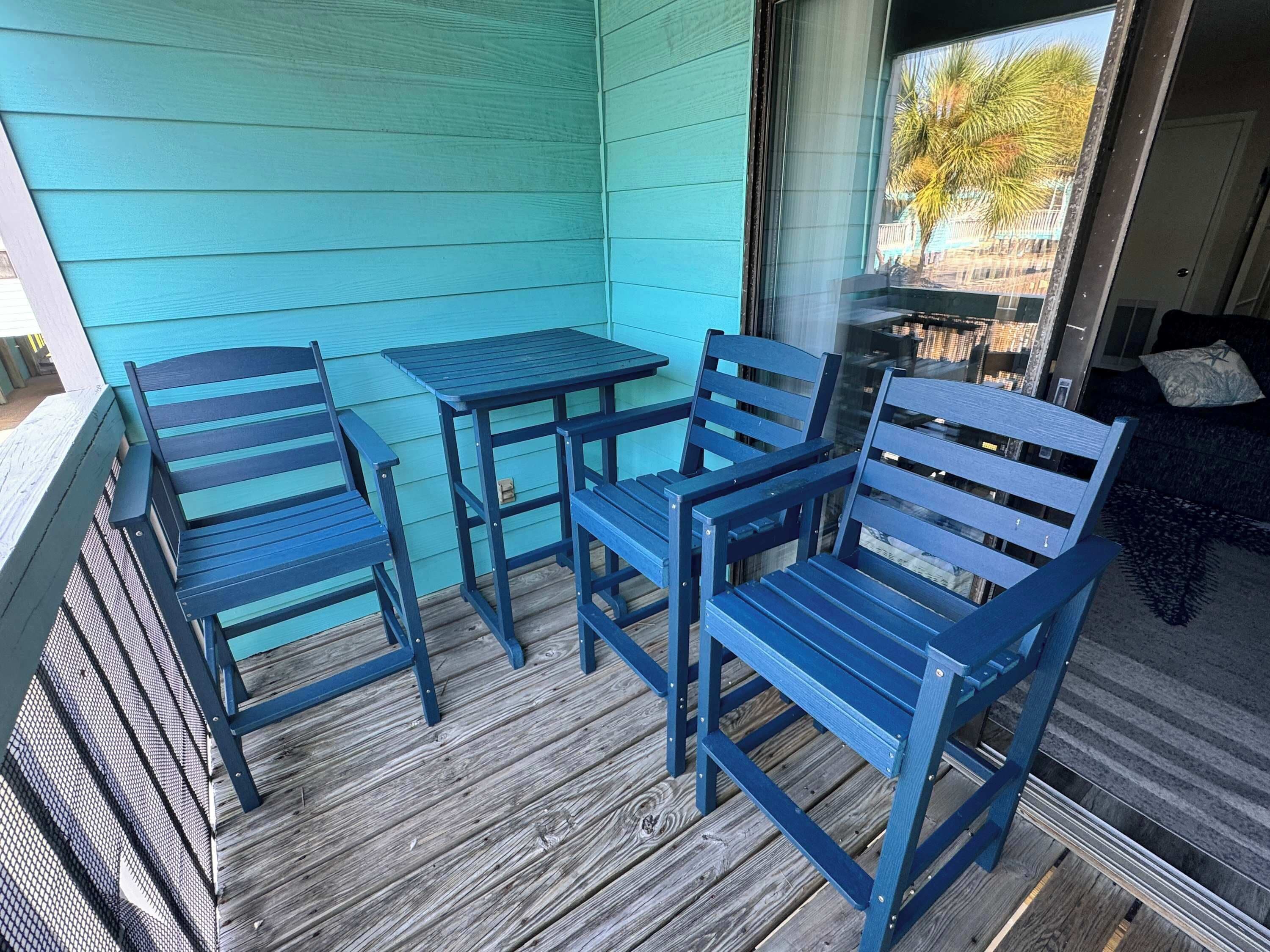 Varanda com mesa e cadeiras azuis, piso de madeira e vista para o exterior.