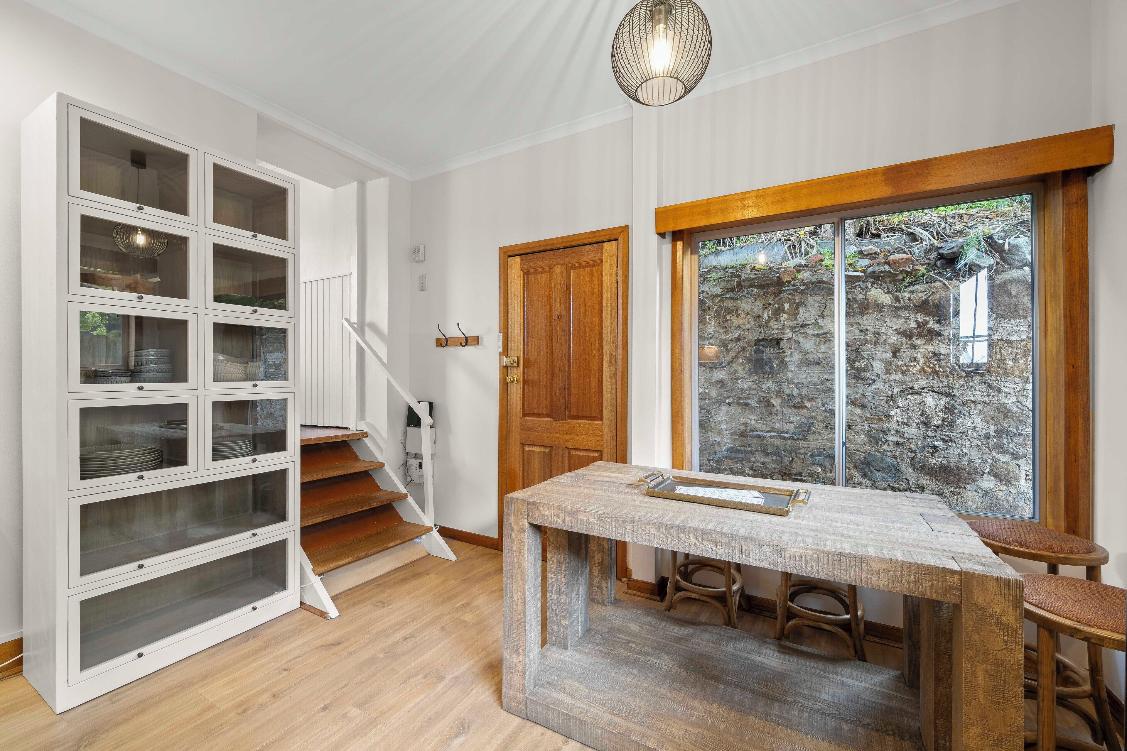 A staircase with a wooden door, a dining area with a rustic table, and a view of a stone wall through a large window.