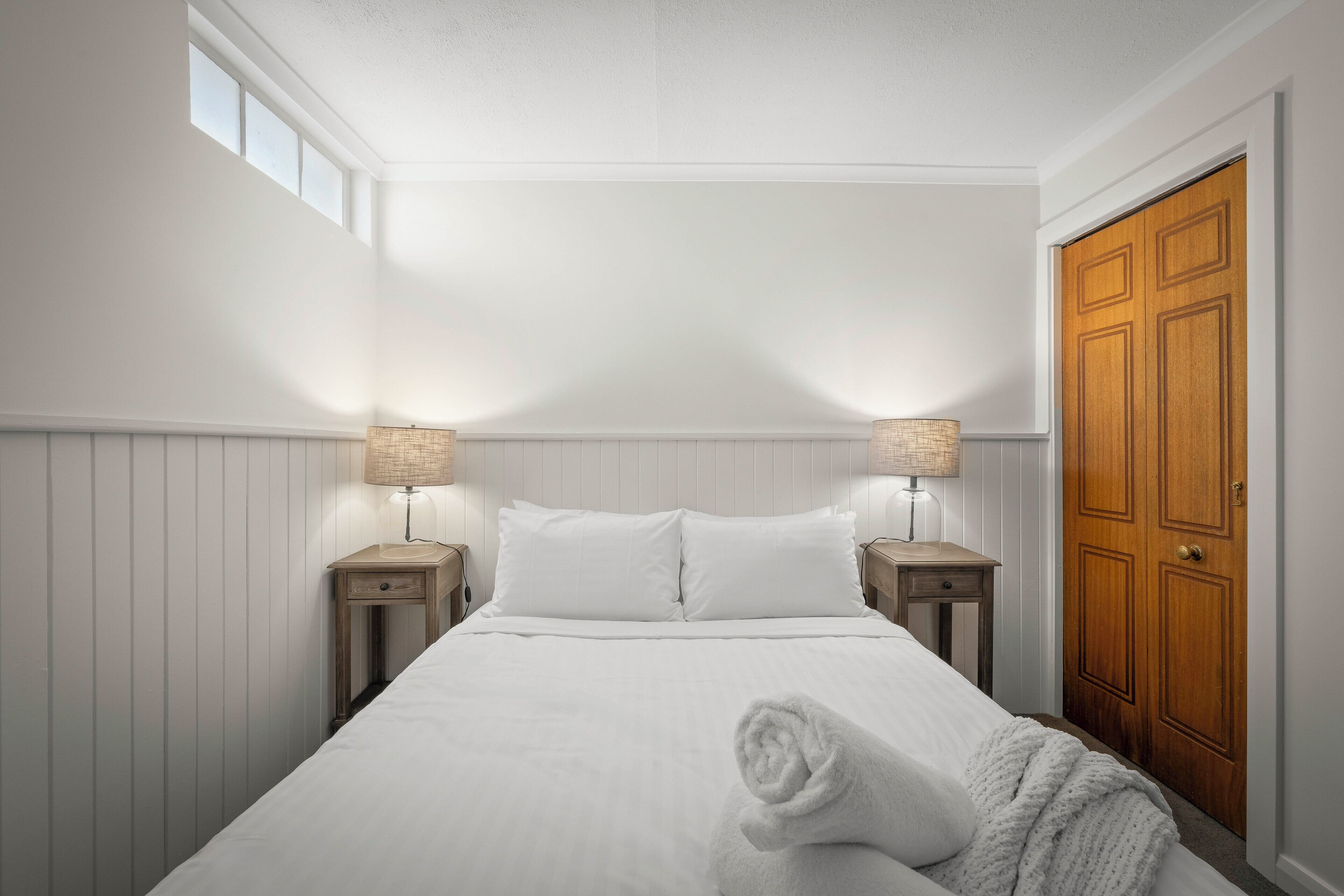 Una cama bien hecha con sábanas blancas, dos mesitas de noche con lámparas, un armario de madera y una ventana que proporciona luz natural.