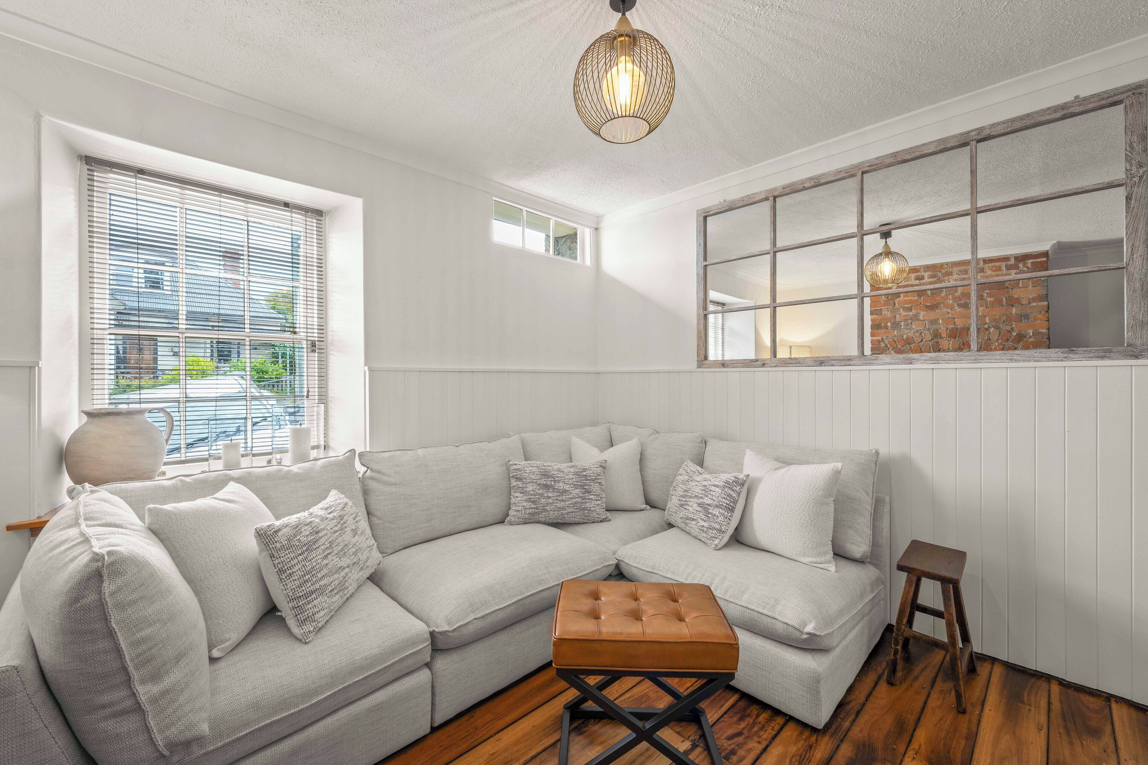 Un salon moderne avec un canapé d’angle, un plancher en bois et une grande fenêtre équipée de stores.