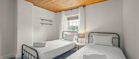 Un dormitorio con dos camas individuales, techo de madera, ventana con cortinas y una mesita de noche con lámpara.