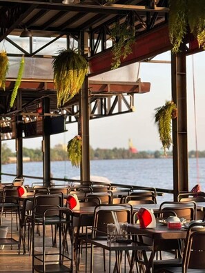 Een buitenterras met tafels en stoelen, hangplanten en uitzicht op het water.