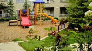 A playground with a slide and climbing structures, surrounded by a gravel area and greenery.