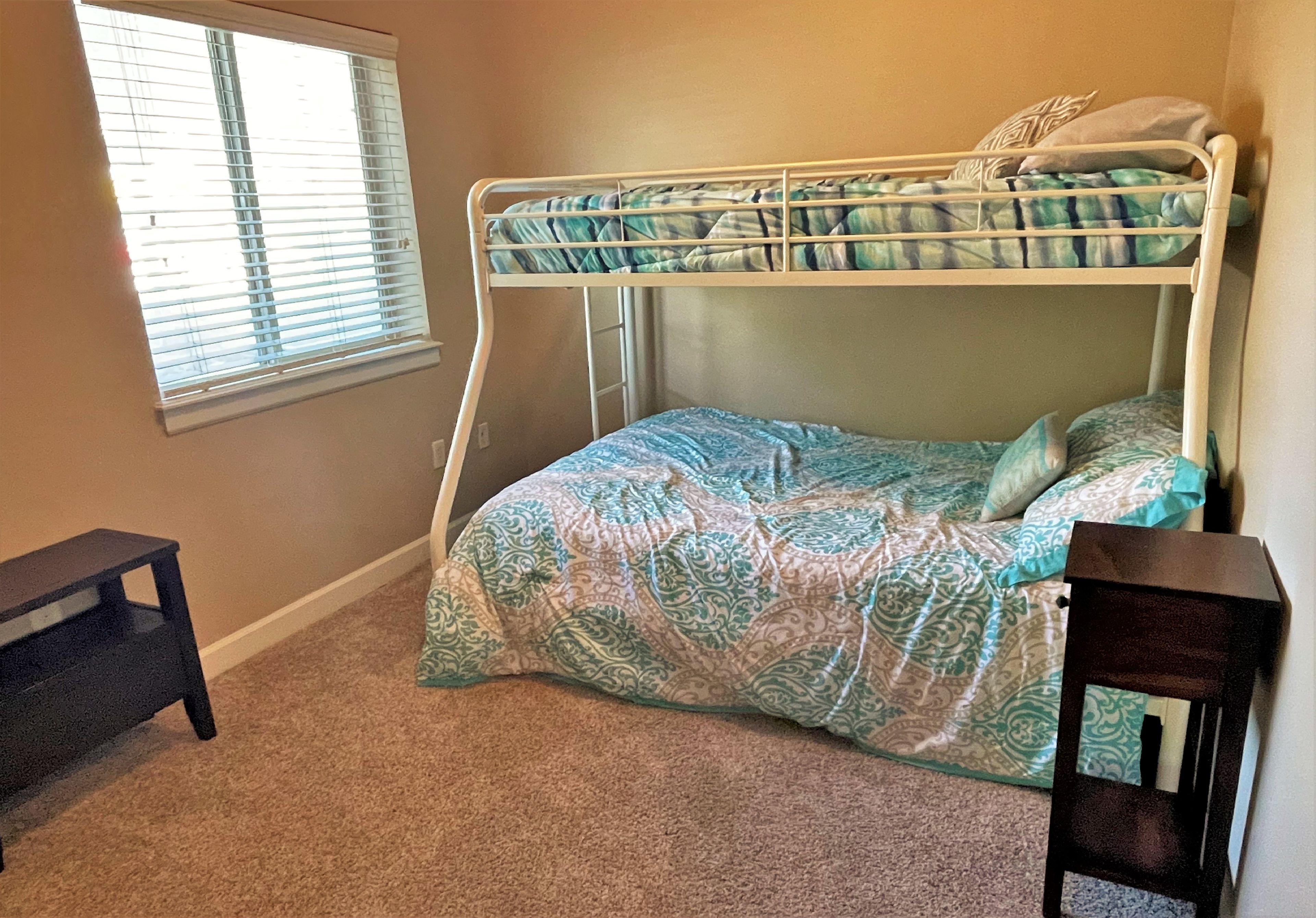 A bunk bed with a patterned bedspread, a small wooden table, and a window with blinds.