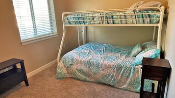 A bunk bed with a patterned bedspread, a small wooden table, and a window with blinds.