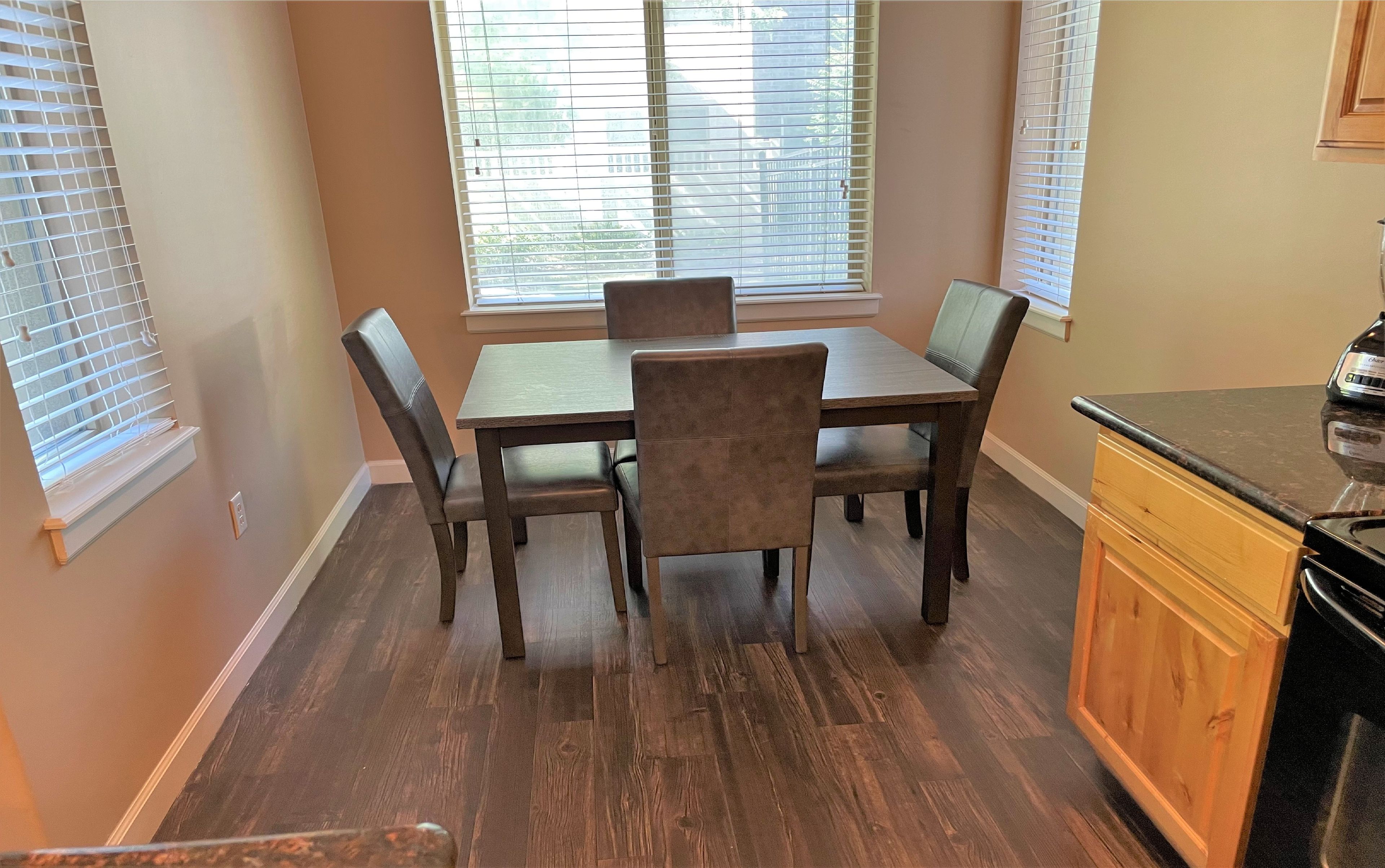 Un coin repas avec une table et des chaises en bois, une fenêtre avec des stores et un comptoir de cuisine.