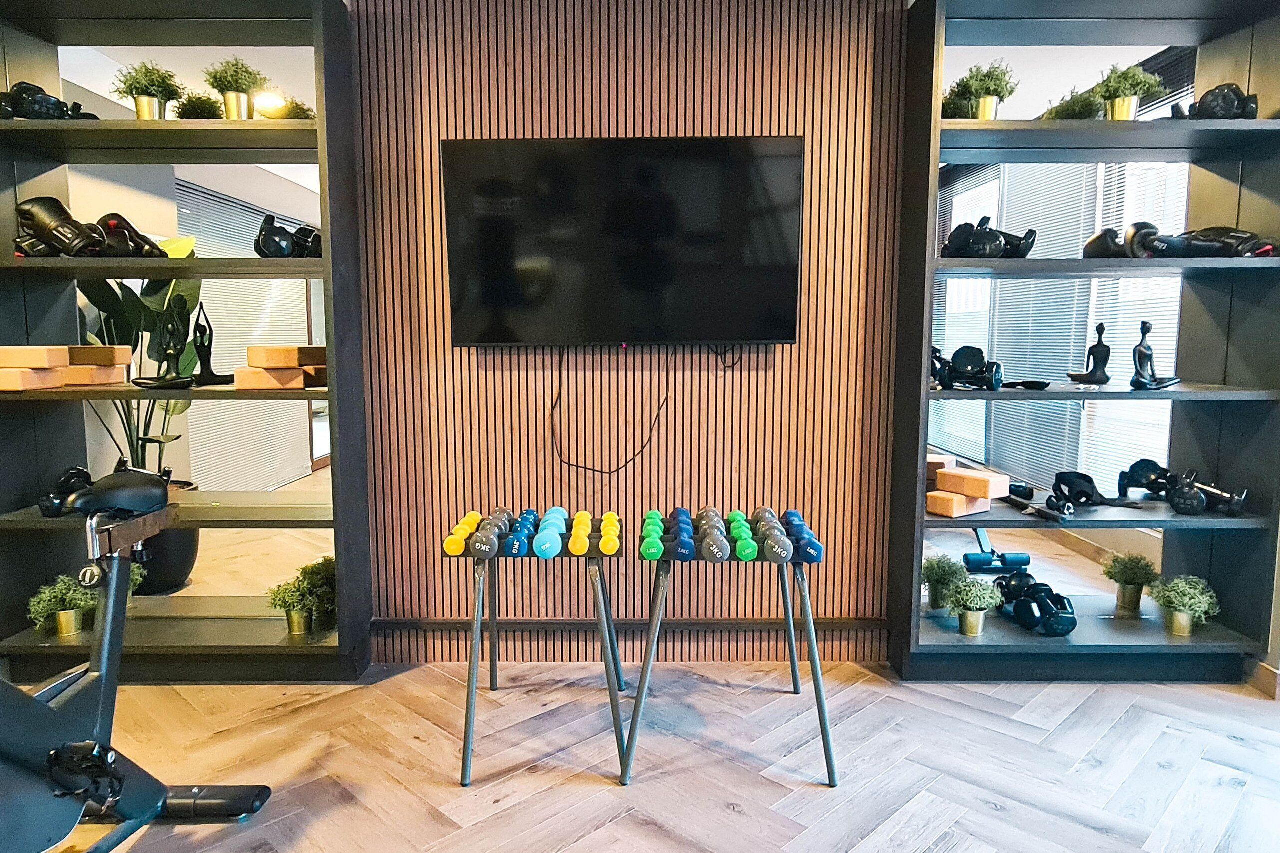 A room with a TV mounted on a wooden wall, shelves with shoes, a dumbbell rack, and a treadmill.
