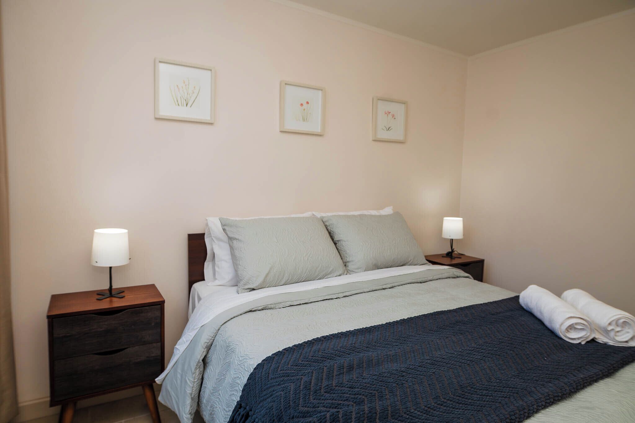 Uma cama bem arrumada com um cobertor azul, duas mesas de cabeceira com abajures e três quadros na parede.