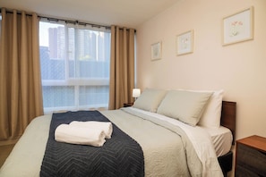 Habitación de hotel con una cama, una ventana con cortinas, una mesita de noche y dos cuadros en la pared.
