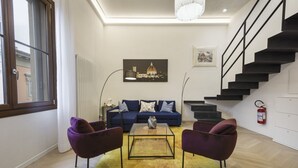 Una sala de estar moderna con un sofá azul, dos sillones morados, una mesa de centro de cristal y una escalera.