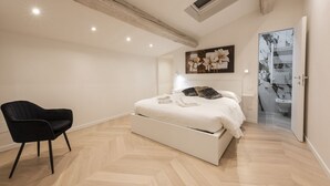 Un dormitorio moderno con una cama, una silla negra y un baño visible a través de una puerta abierta.