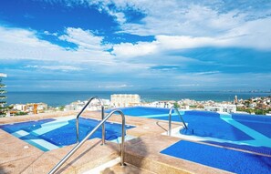 A large outdoor swimming pool with multiple lanes, surrounded by a stone deck and metal railings, overlooking a coastal cityscape.