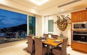 Uma sala de jantar moderna com uma mesa grande e cadeiras de vime, uma peça decorativa na parede e vista para a cidade ao entardecer.