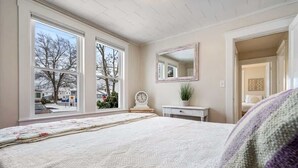 A bedroom with a large bed, a dresser, a mirror, and a window with a view of trees and houses.