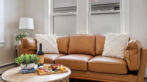 A brown leather sofa with white cushions, a round wooden coffee table with a plant, a bottle of wine, and a board of snacks.
