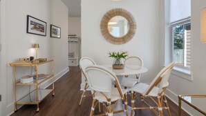 A dining area with a round table, white wicker chairs, a gold-framed mirror, and a window with a view of trees. -  Stylish Downtown Cottage w/ Golf Cart for Rent (Ocean Springs)