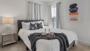 A neatly arranged bedroom with a bed, bedside table, lamp, and a tray with bottles and glasses. -  Stylish Downtown Cottage w/ Golf Cart for Rent (Ocean Springs)